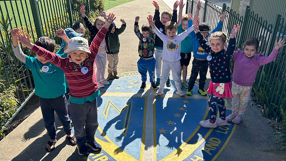 Prep students standing together on a school pathway with arms raised, highlighting joy, belonging and a welcoming early years learning experience at Queen of Apostles Primary School